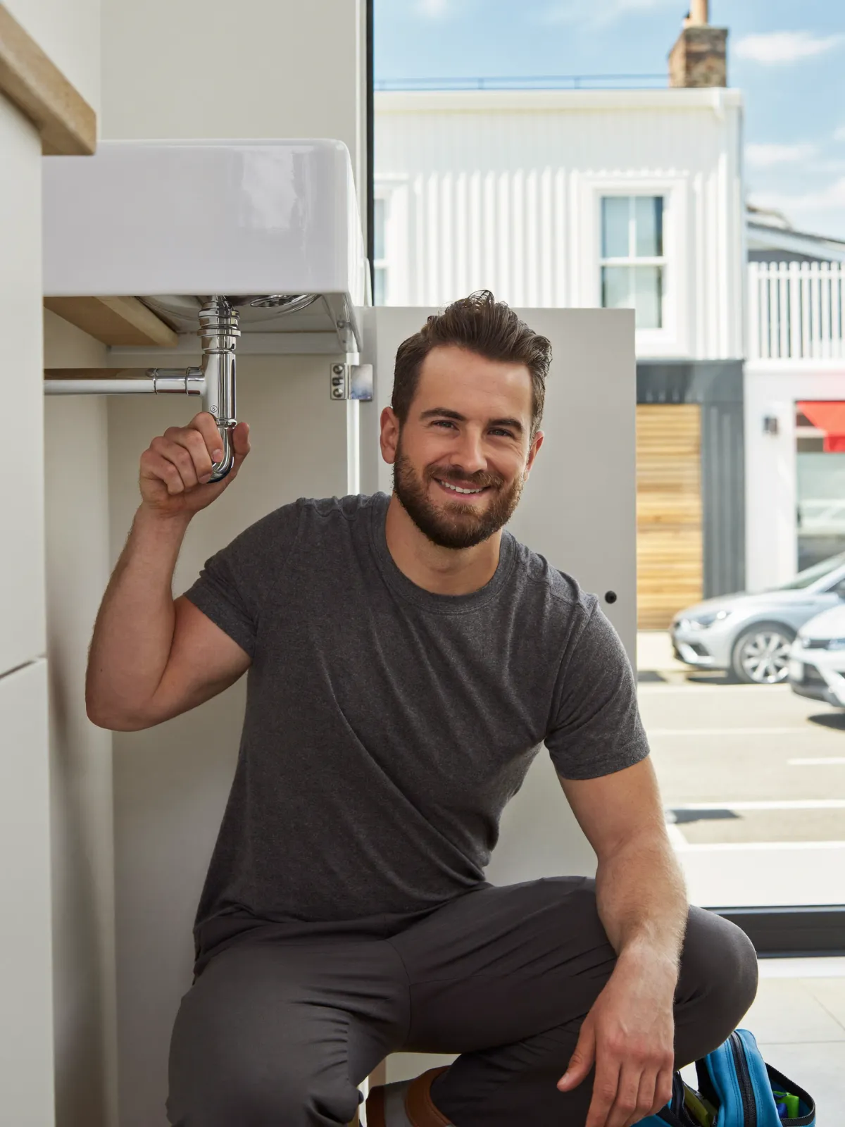 A smiling plumber kneeling by a kitchen sink he is fitting