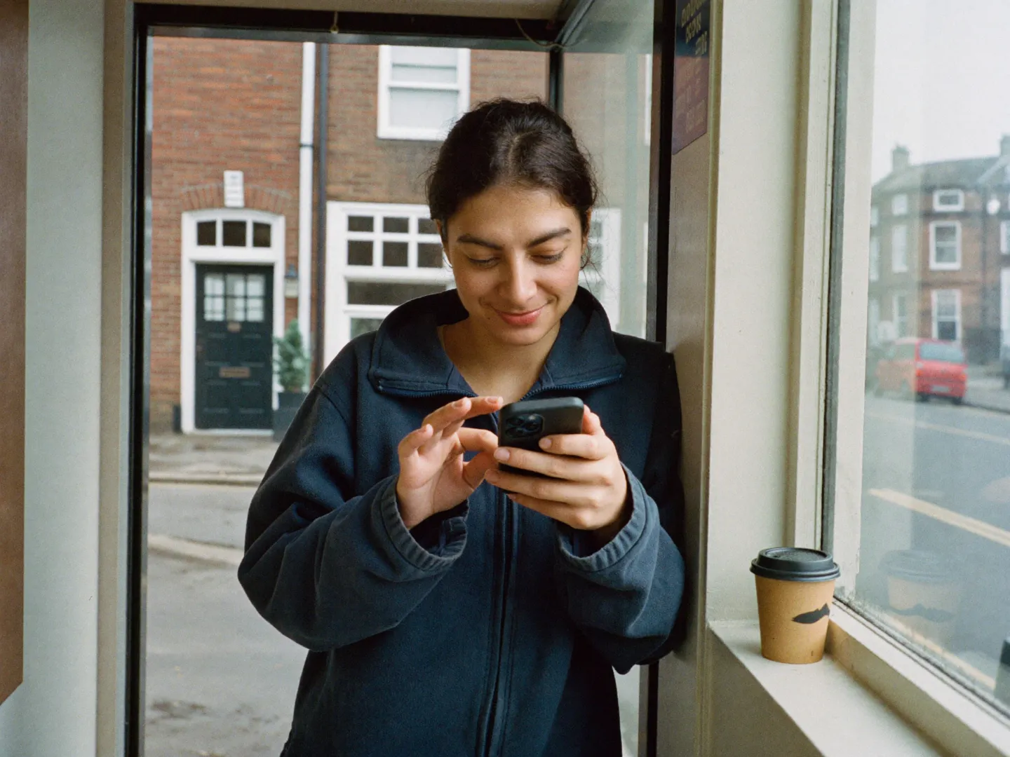 A small business owner smiling as she types a text message in her shop doorway, a takeaway coffee on the window ledge